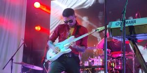 Davide Lo Surdo performing live under red stage lights, wearing sunglasses and a red shirt while playing a white semi-hollow electric guitar.