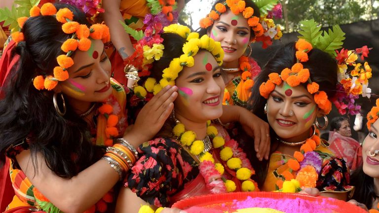 Group of women wearing floral crowns and traditional jewelry celebrating Pahela Falgun with colorful face powder.