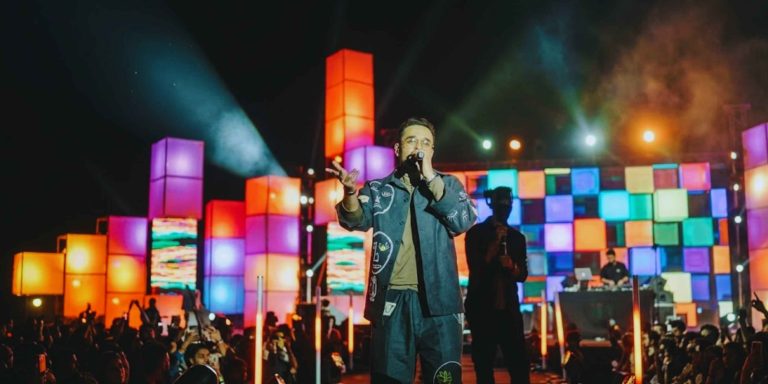 Alemam Elghandour performing live on a grand outdoor stage featuring vibrant, multi-colored cube lighting and a massive crowd during a music festival.