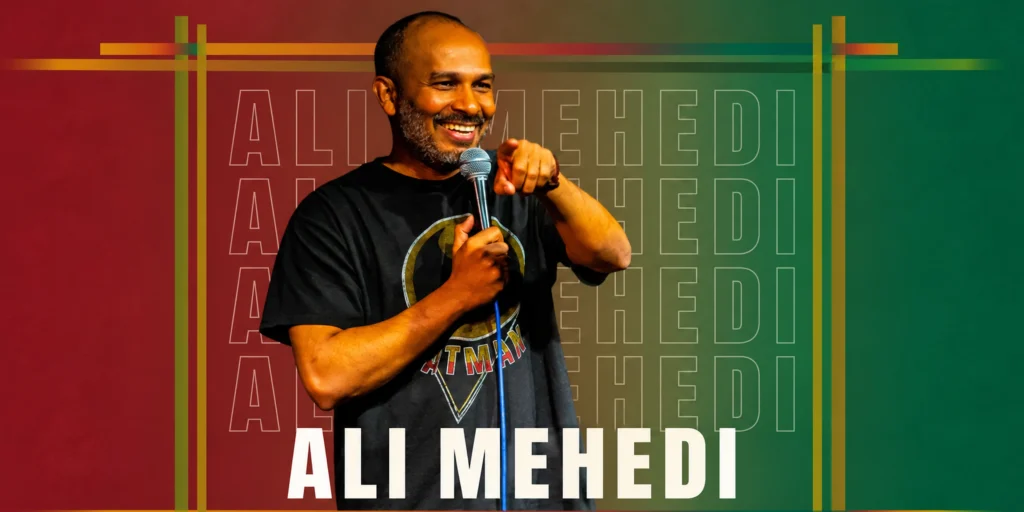 Comedian Ali Mehedi smiling and holding a microphone while performing on stage at a South Asian comedy festival in NYC.