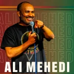 Comedian Ali Mehedi smiling and holding a microphone while performing on stage at a South Asian comedy festival in NYC.